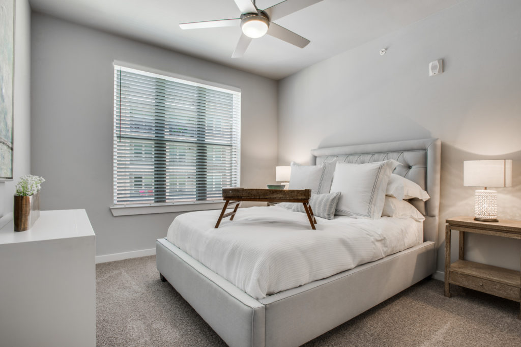 Model apartment bedroom with king bed, large window with wood blinds, and ceiling fan
