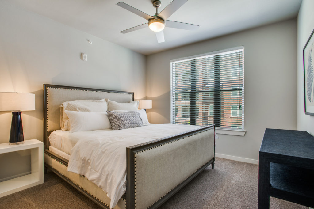 Model apartment bedroom with large bed, ceiling fan, and window with wood blinds