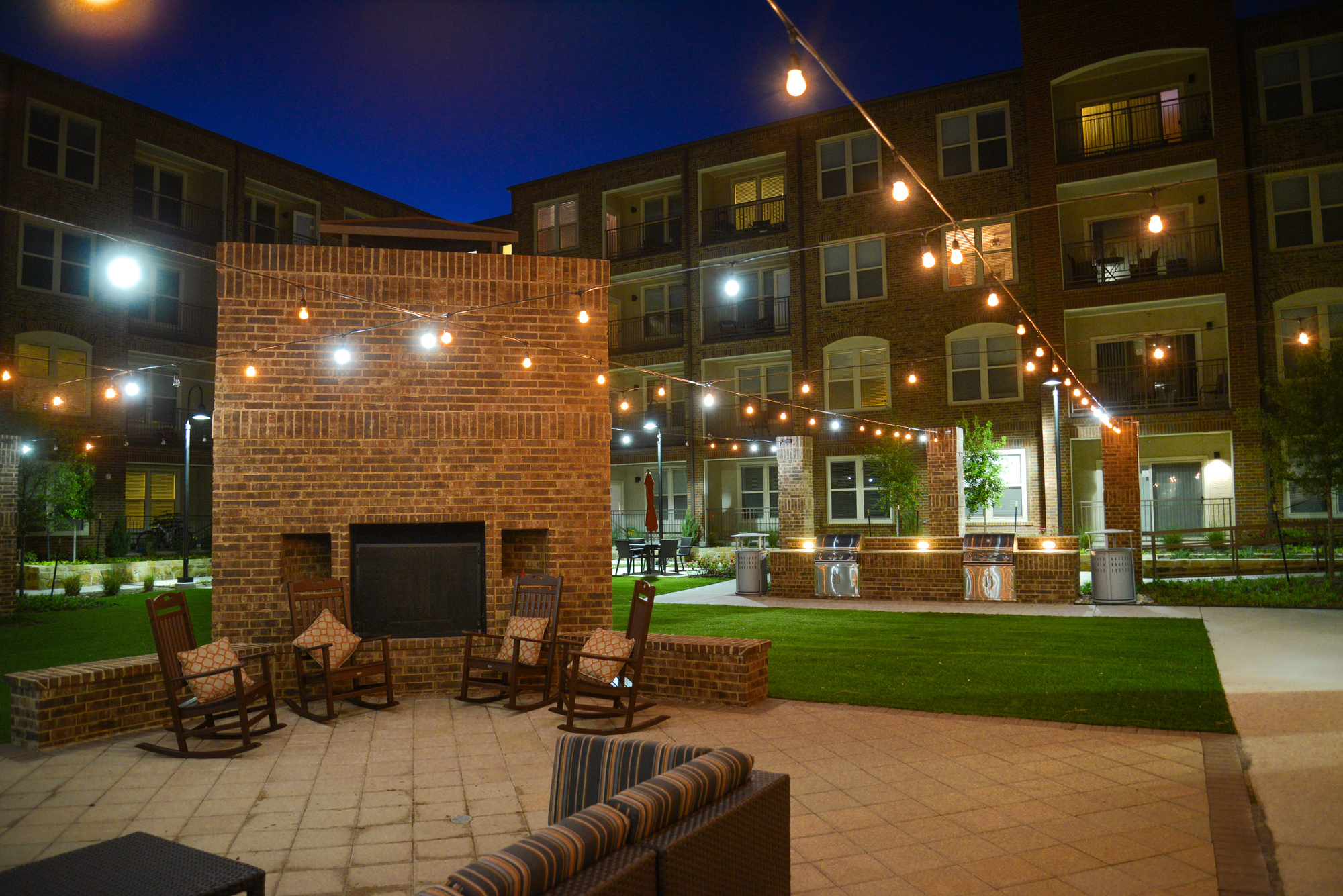 Community courtyard and outdoor kitchen with gas grills and rocking chairs near firepit with festival lights