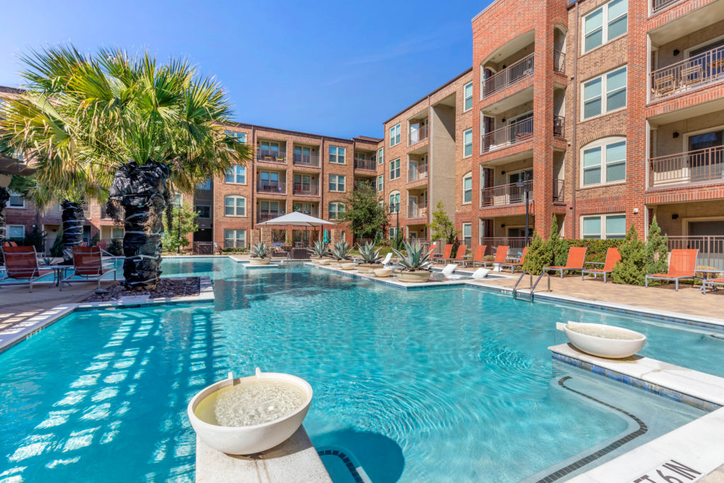 Wide view of swimming pool with water features, palm trees, shaded cabana, lounge seating and apartment balconies in the background.