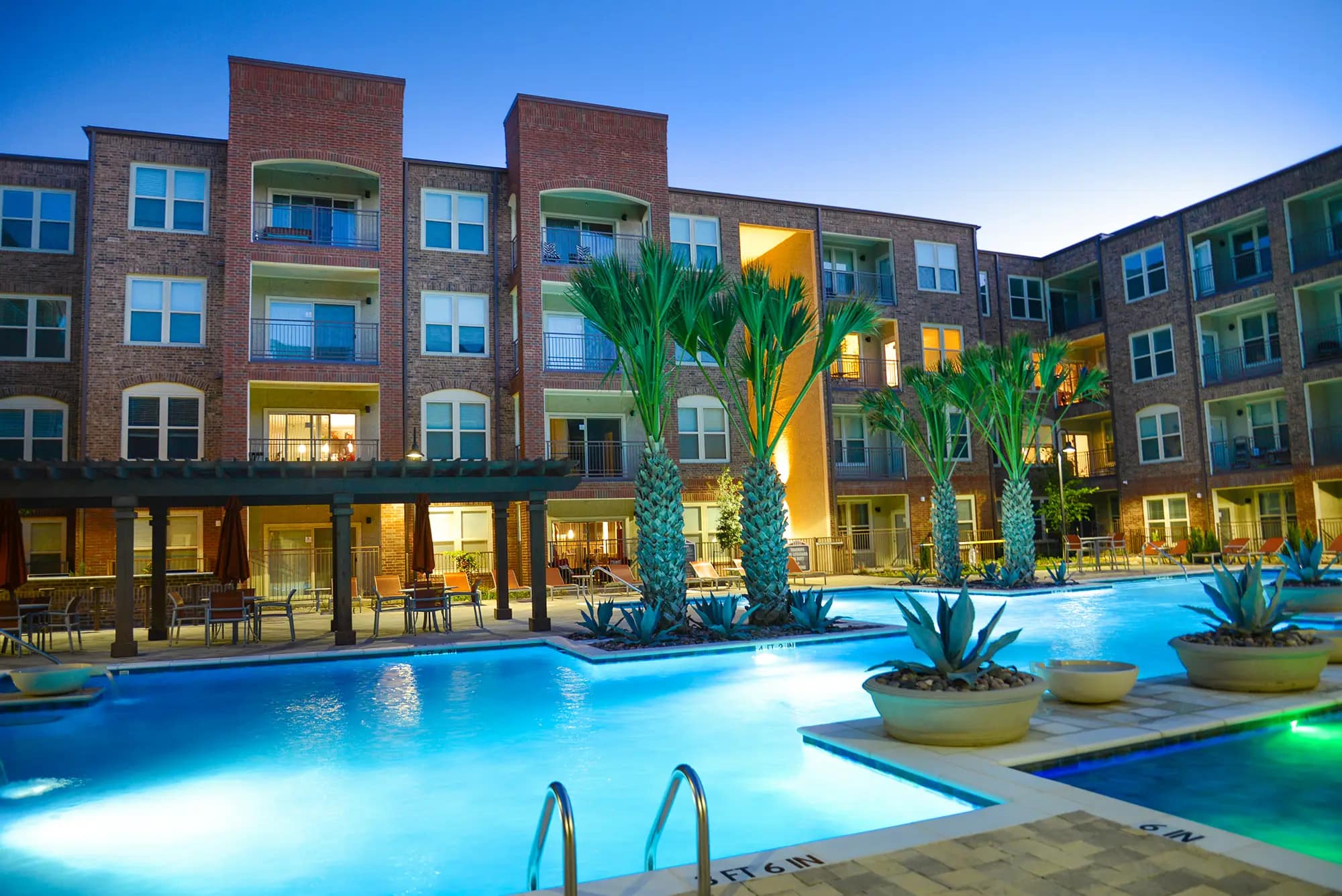 Wide view of swimming pool with water features, palm trees, shaded cabana, lounge seating and apartment balconies in the background.